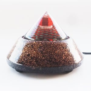 May include: A clear glass pyramid with a red and green geometric pattern inside sits on a brown, textured base. The base has a black rim and two small black feet.