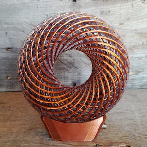 Pode incluir: Escultura decorativa com design espiralado e centro aberto. Feita de metal cor de cobre, apresenta um padrão repetitivo de pequenos elementos quadrados. A base geométrica também é de cobre. O efeito geral é uma peça artística moderna.