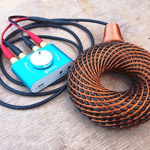 Pode incluir: Um receptor de áudio Bluetooth turquesa com um botão de volume prateado e cabos pretos conectados a um alto-falante toroidal de cor cobre com uma textura de treliça trançada e um acessório superior liso de cor cobre.