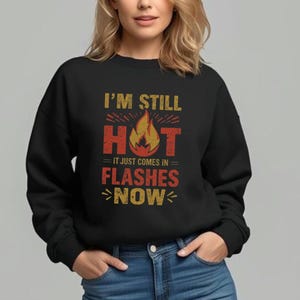 Puede incluir: Sudadera negra con el texto "I'M STILL HOT it just comes in FLASHES NOW" en dorado y rojo. El gráfico incluye un diseño de llama. La modelo lleva vaqueros azules.