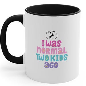 Peut inclure: Une tasse à café en céramique blanche avec un bord noir. La tasse porte une inscription amusante qui dit "J'étais normal il y a deux enfants". Le texte est en rose, bleu et jaune. La tasse a deux yeux de dessin animé au-dessus du texte.