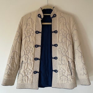 Pode incluir: Casaco acolchoado creme com bordado floral e forro azul marinho. O casaco apresenta gola mandarim, fechos de rã e um design reversível. O bordado é em cor azul escuro contrastante.