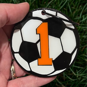 May include: A round soccer ball ornament with a black and white pattern and the number 1 in orange. The ornament has a black string for hanging and is held against a background of green grass.