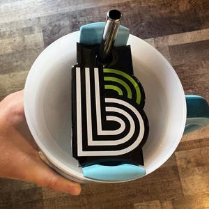 May include: White ceramic mug with a teal handle and a black and white graphic. The graphic is a stylized letter "B" with green accents. A stainless steel straw is inserted into the mug.