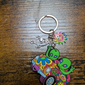 K&ouml;nnte beinhalten: Ein Schl&uuml;sselanh&auml;nger mit mehreren Anh&auml;ngern. Der Hauptanh&auml;nger zeigt zwei gr&uuml;ne Aliens in einem bunten Van mit dem Wort "Peace". Weitere Anh&auml;nger sind eine Rakete und eine Sonne im psychedelischen Design.