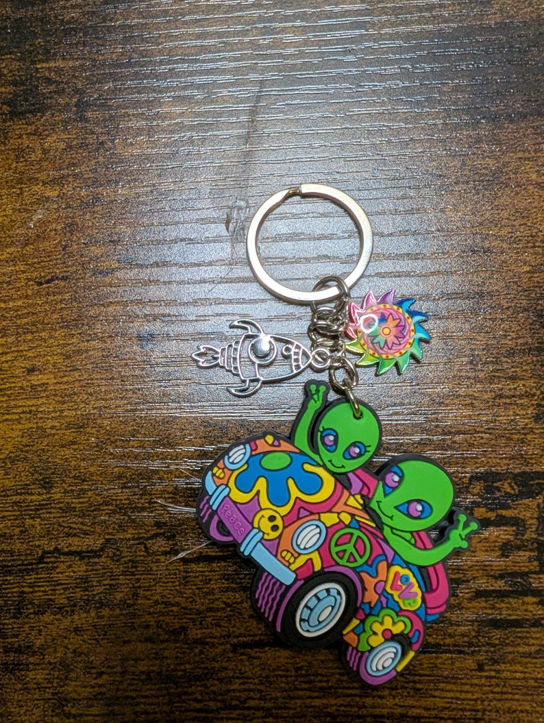 K&ouml;nnte beinhalten: Ein Schl&uuml;sselanh&auml;nger mit mehreren Anh&auml;ngern. Der Hauptanh&auml;nger zeigt zwei gr&uuml;ne Aliens in einem bunten Van mit dem Wort "Peace". Weitere Anh&auml;nger sind eine Rakete und eine Sonne im psychedelischen Design.