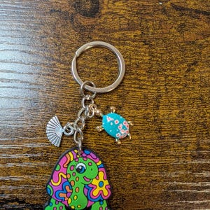 K&ouml;nnte beinhalten: Ein silberner Schl&uuml;sselanh&auml;nger mit drei Anh&auml;ngern: eine gr&uuml;ne Schildkr&ouml;te mit bunten Wirbeln, ein blauer Marienk&auml;fer mit rosa Blumen und ein silberner F&auml;cher. Der Schildkr&ouml;tenanh&auml;nger ist der gr&ouml;&szlig;te und prominenteste.