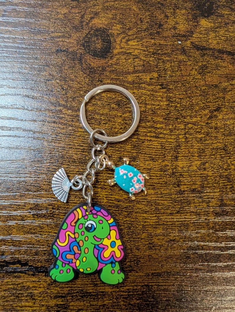 K&ouml;nnte beinhalten: Ein silberner Schl&uuml;sselanh&auml;nger mit drei Anh&auml;ngern: eine gr&uuml;ne Schildkr&ouml;te mit bunten Wirbeln, ein blauer Marienk&auml;fer mit rosa Blumen und ein silberner F&auml;cher. Der Schildkr&ouml;tenanh&auml;nger ist der gr&ouml;&szlig;te und prominenteste.