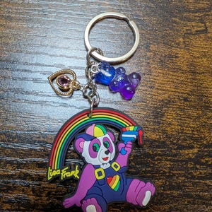 K&ouml;nnte beinhalten: Ein Lisa Frank Schl&uuml;sselanh&auml;nger mit einem lila Pandab&auml;ren, einem Regenbogen und einer Wasserpistole. Der Schl&uuml;sselanh&auml;nger enth&auml;lt einen goldenen Herzanh&auml;nger mit einem lila Edelstein und zwei Gummib&auml;rchen-Anh&auml;ngern. Der Text "Lisa Frank" ist sichtbar.