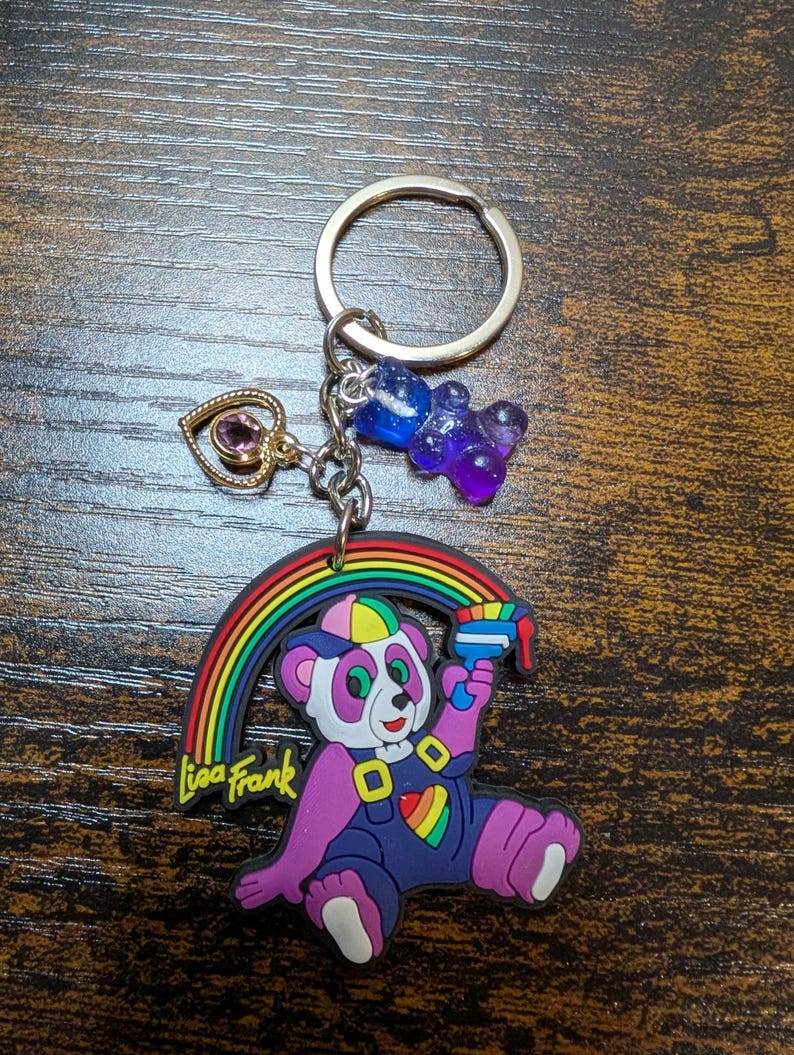 K&ouml;nnte beinhalten: Ein Lisa Frank Schl&uuml;sselanh&auml;nger mit einem lila Pandab&auml;ren, einem Regenbogen und einer Wasserpistole. Der Schl&uuml;sselanh&auml;nger enth&auml;lt einen goldenen Herzanh&auml;nger mit einem lila Edelstein und zwei Gummib&auml;rchen-Anh&auml;ngern. Der Text "Lisa Frank" ist sichtbar.