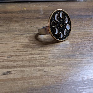 Pode incluir: Um anel de prata com um design de fases lunares em preto e branco. O anel está sobre uma superfície de madeira.