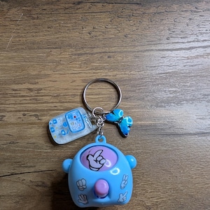 Puede incluir: Un llavero azul y rosa con un pequeño amuleto de teléfono móvil, un amuleto de mariposa y un juego de manos giratorias. El juego tiene un botón rosa y cuatro gestos de manos diferentes alrededor del botón.