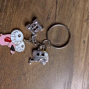 Op de afbeelding: Een zilveren sleutelhanger met een roze cartoonhond met grote ogen, een zilveren camper en een zilveren verrekijker.