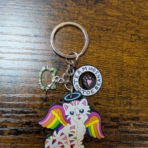 K&ouml;nnte beinhalten: Ein silberner Schl&uuml;sselanh&auml;nger mit einem Anh&auml;nger, der eine wei&szlig;e Katze mit Regenbogenfl&uuml;geln und einem Heiligenschein zeigt. Der Schl&uuml;sselanh&auml;nger enth&auml;lt auch einen Anh&auml;nger mit dem Text "TEAM ODDS BEAN TOE". Ein kleiner gr&uuml;ner Charm mit Strasssteinen ist ebenfalls angebracht.