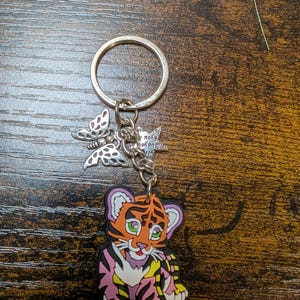 K&ouml;nnte beinhalten: Ein Schl&uuml;sselanh&auml;nger mit einem Cartoon-Tigerbaby-Anh&auml;nger mit orangefarbenen, rosa und gelben Streifen. Der Schl&uuml;sselanh&auml;nger enth&auml;lt einen silbernen Ring, einen Schmetterlingsanh&auml;nger und einen kleinen Anh&auml;nger mit Text.