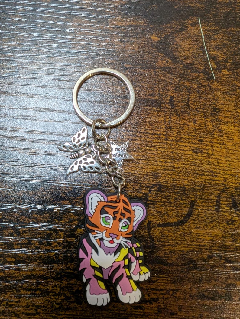 K&ouml;nnte beinhalten: Ein Schl&uuml;sselanh&auml;nger mit einem Cartoon-Tigerbaby-Anh&auml;nger mit orangefarbenen, rosa und gelben Streifen. Der Schl&uuml;sselanh&auml;nger enth&auml;lt einen silbernen Ring, einen Schmetterlingsanh&auml;nger und einen kleinen Anh&auml;nger mit Text.