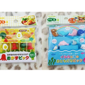 Puede incluir: Dos paquetes de pinchos para comida coloridos. El primer paquete contiene ocho pinchos con dise&ntilde;os de pulpo, huevo, br&oacute;coli y camar&oacute;n. El segundo paquete contiene ocho pinchos con dise&ntilde;os de ping&uuml;ino, foca, delf&iacute;n y oso. El texto en el primer paquete dice "Okazu Bick" y el texto en el segundo paquete dice "Iruka to Umi no Otomo Dachi Pick".