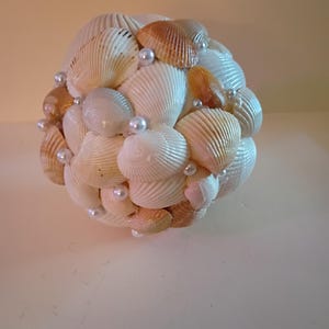 May include: A spherical decorative object crafted from various seashells in shades of white, cream, and tan. Small, round, pearl-like beads are interspersed among the shells. The arrangement creates a textured, natural aesthetic, suitable for coastal decor.