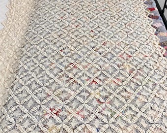 May include: A cream-colored crocheted blanket featuring a repeating diamond pattern with intricate openwork stitching. The textile is draped over a bed, partially revealing a multi-colored patchwork quilt underneath with a scalloped edge finish.