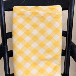 May include: A folded yellow and white checkered fabric, possibly a tablecloth or sheet, rests on a black chair. The fabric's pattern is a classic gingham design. The chair has a woven seat and a decorative back.