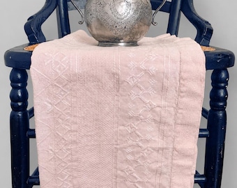 May include: A pale pink rectangular textile featuring intricate white embroidered geometric patterns and a delicate hemstitch border, draped over a dark navy blue wooden chair with a silver metal bowl with two handles placed on top.