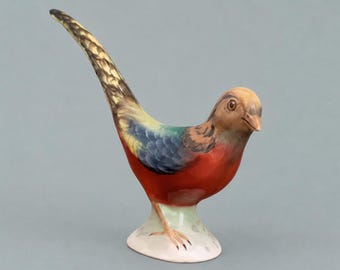 May include: A ceramic bird figurine featuring a vibrant red chest, a blue and yellow patterned wing, and a long, slender tail with brown and black markings. The bird stands on a small, pale base, with a light brown head and a sharp, pointed beak.