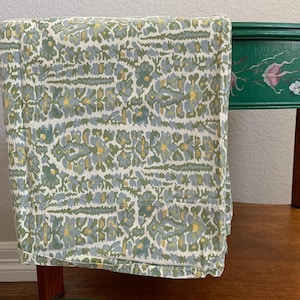 May include: A folded floral patterned fabric in shades of green, blue, and yellow rests on a wooden side table. The table has a dark green painted top with floral accents and a natural wood base.