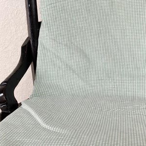 May include: A folded, light green and white checkered fabric draped over a dark wooden chair. The fabric has a small gingham pattern. The chair has a curved back and ornate details. The fabric appears to be a tablecloth or a similar textile.