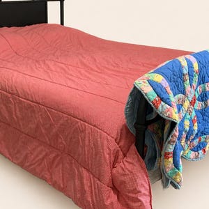 May include: A black metal bed frame with a red and white patterned comforter. A blue and multi-colored quilt is draped over the foot of the bed. The bed is set against a plain background.