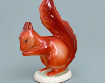 May include: A glossy ceramic figurine of a squirrel in a vibrant orange-red hue. The squirrel is depicted in a seated position with its bushy tail curved upward behind its back. It stands on a small, smooth, off-white oval base.