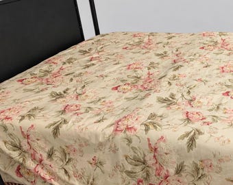 May include: A bedspread featuring a cream-colored base with a repeating floral pattern in shades of pink and muted green. The fabric is draped over a black metal bed frame, showcasing a decorative twisted rope trim along the bottom edge of the cover.
