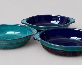 May include: Three round ceramic baking dishes with side handles. One dish is a vibrant turquoise color, while the other two feature a deep navy blue interior with a turquoise rim and a dark, textured brown exterior finish on the base.
