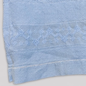 May include: A light blue textile featuring intricate embroidery with geometric patterns, including diamond shapes and cross-stitch details. The fabric has a textured weave and a decorative border with a delicate hemstitch along the bottom edge.
