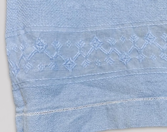 May include: A light blue textile featuring intricate embroidery with geometric patterns, including diamond shapes and cross-stitch details. The fabric has a textured weave and a decorative border with a delicate hemstitch along the bottom edge.