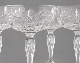 May include: A set of four clear crystal coupe glasses featuring intricate etched leaf patterns on the bowls and bases. Each glass has a slender, faceted stem with a vertical ribbed design, providing a sophisticated look for serving beverages.