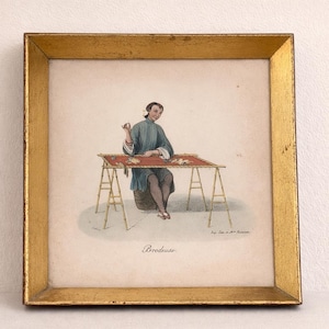 May include: Framed print of a person embroidering at a table. The person is wearing a blue and white outfit. The table has a red top and is supported by a bamboo-like structure. The frame is gold.