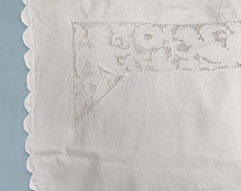 May include: A white rectangular pillowcase featuring a scalloped edge and an intricate cutwork embroidery panel in the corner. The embroidery displays a detailed floral and leaf pattern integrated into a fine mesh grid, crafted from white fabric.