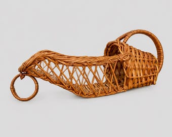 May include: A brown woven wicker wine bottle holder featuring an elongated cradle design with a lattice pattern, a circular ring handle at the front, and a curved handle at the back. The structure is crafted from natural reed material.