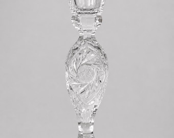 May include: A clear, cut-glass candlestick holder with a square top and a round base. The stem features a detailed, swirling pattern. The candlestick is designed to hold a single candle and is suitable for home decor.