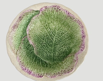 May include: A round ceramic plate featuring a molded cabbage leaf design. The surface displays a textured, veined pattern in shades of vibrant green, with delicate purple-tinted edges and a cream-colored scalloped border surrounding the leaves.