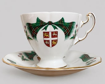 May include: A white porcelain teacup and saucer set featuring a green and red tartan ribbon motif. The cup displays a central crest with a white cross on a red shield, flanked by golden lions. The rim and handle are accented with gold trim.