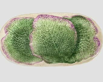 May include: An oval ceramic serving platter featuring a textured, embossed design of three overlapping cabbage leaves. The leaves are colored in shades of vibrant green with distinct purple-tinted edges and a cream-colored base rim.