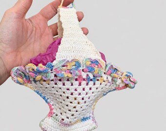 May include: A small, off-white crocheted basket featuring a decorative scalloped rim with pastel-colored accents in pink, blue, yellow, and green. The basket has a single upright handle and contains a crumpled piece of bright magenta tissue paper.