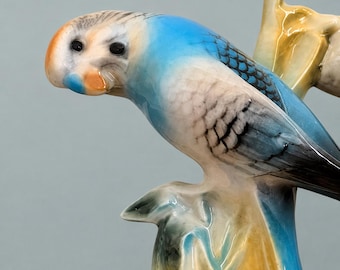 May include: A ceramic figurine of a budgerigar perched on a branch. The bird features a bright blue back and wings with black scalloped markings, a white chest, and a pale orange face with black eyes. The branch is glazed in shades of yellow and brown.
