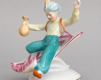May include: A porcelain figurine of a figure wearing a light blue turban with a feather, a pale yellow shirt, and teal trousers, sitting on a flying carpet with a pink and white pattern. The figure holds a tan pouch and is mounted on a white base.