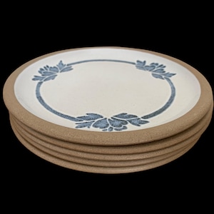 May include: A stack of four round ceramic plates with a speckled cream-colored center and a wide, unglazed tan rim. Each plate features a circular blue floral pattern printed in the middle, creating a consistent design across the entire set.
