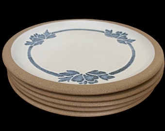 May include: A stack of four round ceramic plates with a speckled cream-colored center and a wide, unglazed tan rim. Each plate features a circular blue floral pattern printed in the middle, creating a consistent design across the entire set.