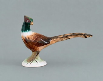 May include: A ceramic pheasant figurine featuring a glossy finish with a dark green head, a white neck band, and brown patterned feathers on the body and long tail. The bird stands on a small, rounded white base with visible dark feet.