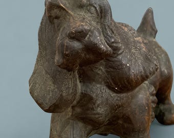 May include: A dark brown, cast metal figurine of a dog with long, drooping ears and a textured surface. The dog is depicted in a standing position with its head slightly tilted, showcasing detailed facial features and a short, upright tail.