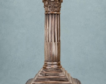 May include: A metallic bronze-toned candle holder shaped like a classical column. It features a square stepped base, a fluted cylindrical shaft, and an ornate Corinthian-style capital with intricate leaf detailing at the top.